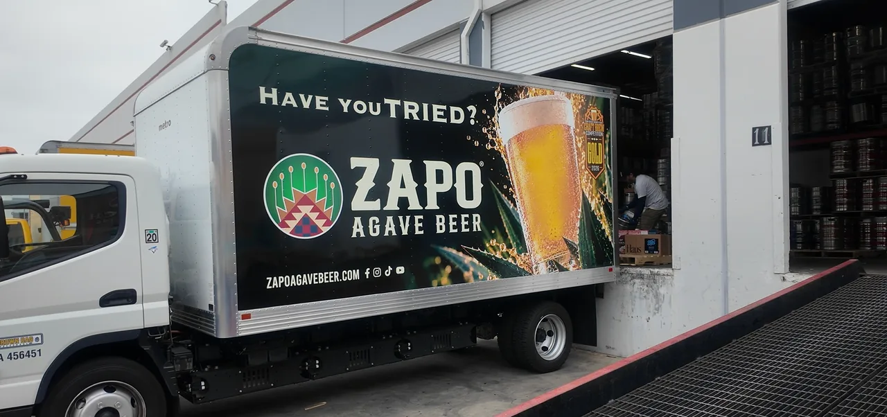 A Brown Bag Beverage RIZON electric box truck with a beer advertisement on its cargo box, backed up to an open loading dock where a worker moves kegs