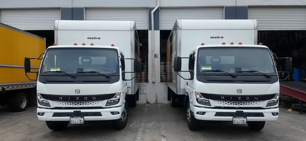 Two white Brown Bag Beverage RIZON electric box trucks parked side by side, facing forward, in front of warehouse loading docks